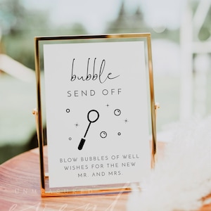 May include: A gold-framed sign with a white background and black text that reads "bubble send off". A black and white illustration of a bubble wand with bubbles is below the text. The text below the illustration reads "Blow bubbles of well wishes for the new Mr. and Mrs."