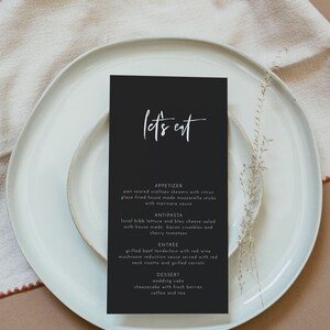 May include: A black menu card with white text that reads "let's eat" on a white plate. The menu lists appetizers, antipasta, entree, and dessert options. The plate is on a beige linen napkin.