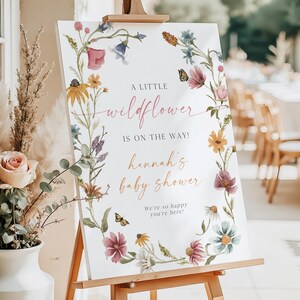 May include: A white sign with a floral wreath border. The sign says "A little wildflower is on the way! Hannah's baby shower. We're so happy you're here!"