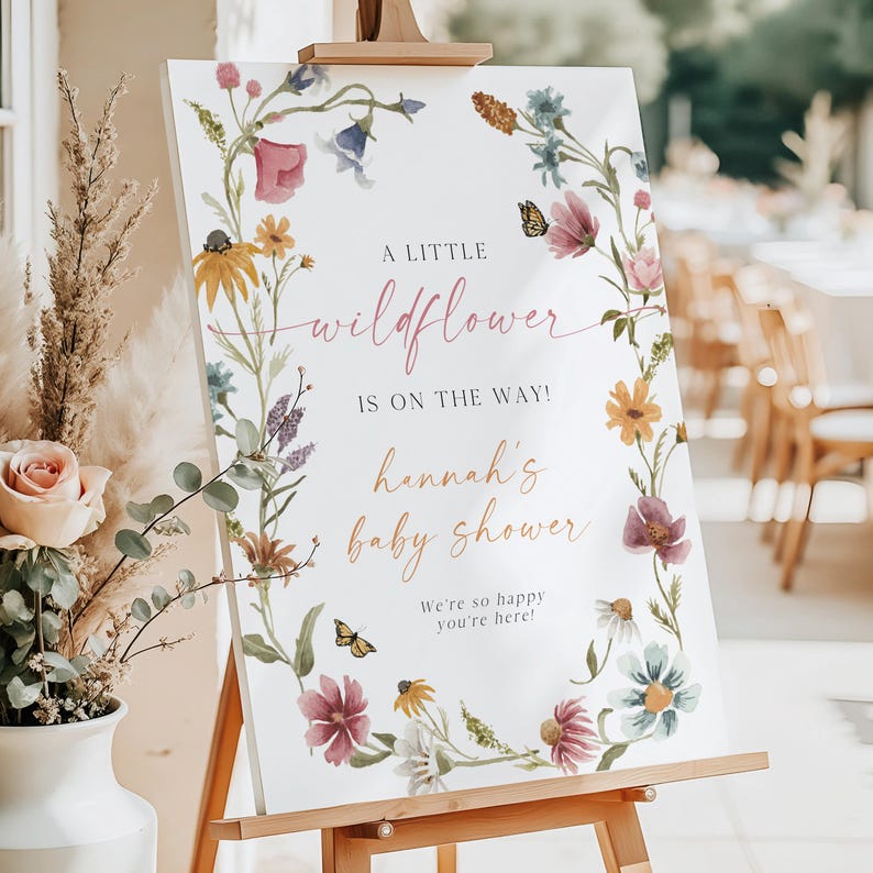 May include: A white sign with a floral wreath border. The sign says "A little wildflower is on the way! Hannah's baby shower. We're so happy you're here!"