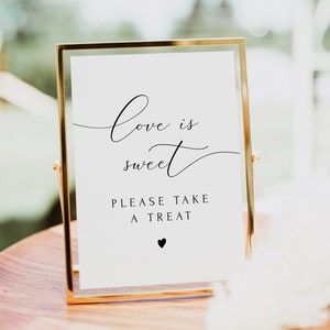 Peut inclure: Panneau doré avec l'inscription "Love is sweet, please take a treat" en noir élégant. Un petit cœur noir en bas. Le panneau est posé sur une surface en bois, idéal pour un mariage ou un événement.