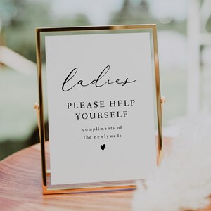 May include: A gold-framed sign with the word "Ladies" in elegant script. Below, it reads "PLEASE HELP YOURSELF" and "compliments of the newlyweds" with a heart symbol. The sign is on a wooden surface.