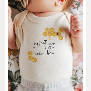 May include: A white baby onesie with a bee and honeycomb design. The onesie says "sweet as can bee".