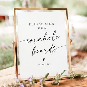 May include: A gold-framed sign with the text "Please sign our cornhole boards" in black script font. A small black heart is below the text, followed by "Thank you!"
