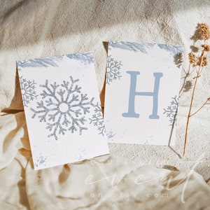 May include: Two white cards with a winter theme. One card features a large, glittery silver snowflake, and the other displays the letter "H" in light blue. Both cards have snowflake and pine branch accents.