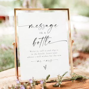 May include: A white sign in a gold frame with black text that reads "message in a bottle". The sign instructs guests to write a message, roll it up, and slip it into a bottle. It also says to leave well wishes for the new Mr. and Mrs.