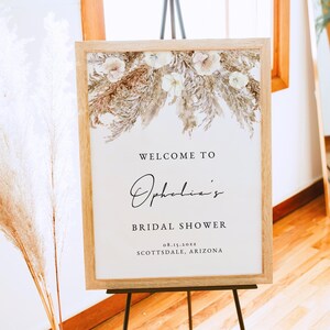 May include: A welcome sign for a bridal shower with a floral design in shades of brown and white. The sign reads "Welcome to Ophelia's Bridal Shower 08.15.20XX Scottsdale, Arizona".