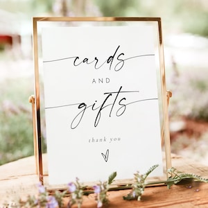 May include: A white sign with black calligraphy that reads "cards and gifts thank you" with a small heart at the bottom. The sign is in a gold frame and is sitting on a wooden table.