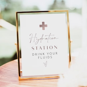 May include: A gold-framed sign with a white background and the text "Hydration Station Drink Your Fluids" in a cursive font. A small pink heart is at the bottom of the sign. A white plus sign is at the top of the sign.