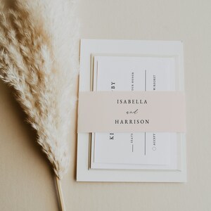 May include: A white wedding invitation with a blush pink ribbon. The ribbon has the text "ISABELLA and HARRISON" printed on it. The invitation also has the text "BY OUR HONOR" and "W/REGRET" printed on it.