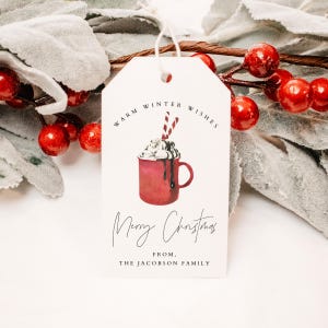 May include: White gift tag with a red and white illustration of a mug of hot chocolate with whipped cream and candy canes. The text on the tag reads "Warm Winter Wishes Merry Christmas From, The Jacobson Family".