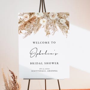 May include: White welcome sign with a floral design featuring dried pampas grass and white flowers. The sign reads "Welcome to Ophelia's Bridal Shower 08.15.20XX Scottsdale, Arizona".