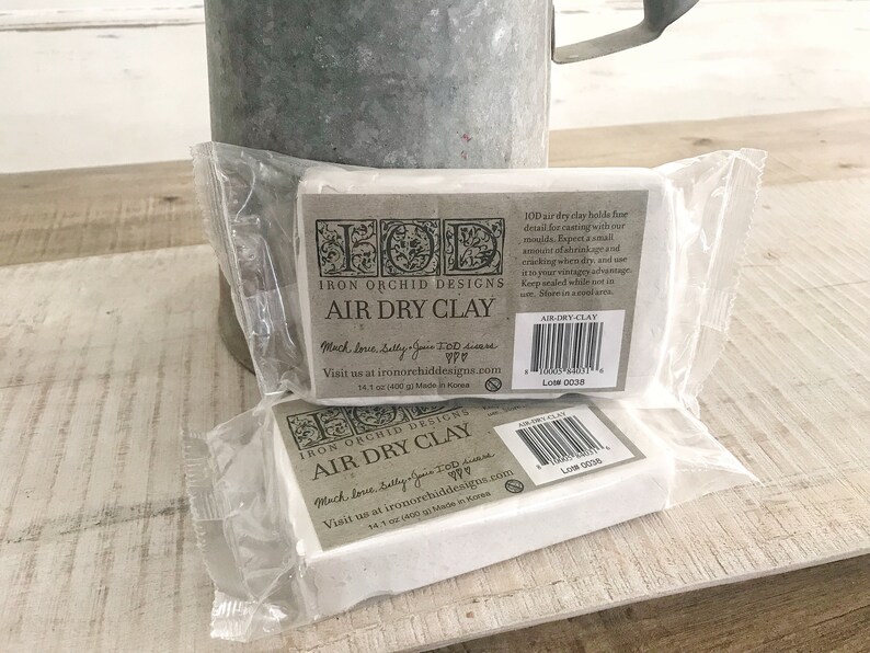 Op de afbeelding: Twee pakketten witte lucht drogende klei van Iron Orchid Designs. De pakketten zijn gelabeld met het bedrijfslogo en de woorden "Air Dry Clay". De tekst "Much love, Sally, Jane & TOD" is afgedrukt op de voorkant van de pakketten. De achterkant van de pakketten bevat instructies voor het gebruik van de klei en een lotnummer.