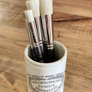 May include: A white ceramic jar with black lettering that reads "BAND MEDAL OF MERIT VIENNA JAMES KEILLER & SON'S DUNDEE MARMALADE PRIZE MEDAL FOR LONDON 1862" holds a set of four paint brushes with white bristles.