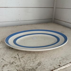 May include: An oval-shaped, white ceramic platter with two concentric blue stripes around the rim. The plate is set on a distressed, white-painted wooden surface.