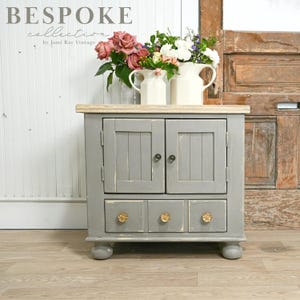 May include: A gray painted wooden cabinet with two doors and three drawers. The cabinet has a distressed finish and gold knobs. The cabinet is sitting on a wooden floor.