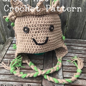 Puede incluir: Patrón de gorro Groot tejido a ganchillo en color beige y verde. El gorro presenta una cara sonriente con ojos de botón y una boca cosida en negro. Tiene orejeras con lazos trenzados y detalles verdes. El texto "Groot Hat Crochet Pattern" es visible.