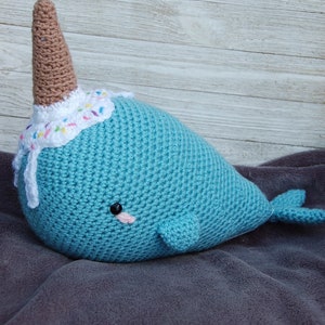 May include: A crocheted narwhal plush toy with a brown horn and a white and colorful frosting hat. The narwhal is laying on a gray blanket.