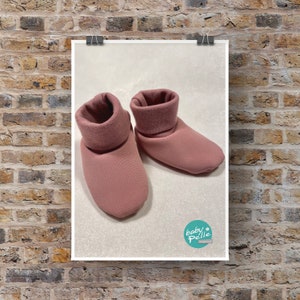 Könnte beinhalten: Ein Paar rosa Baby-Schühchen mit umgeschlagenem Bündchen. Die Schühchen sind aus weichem, plüschem Stoff und haben ein schlichtes Design.