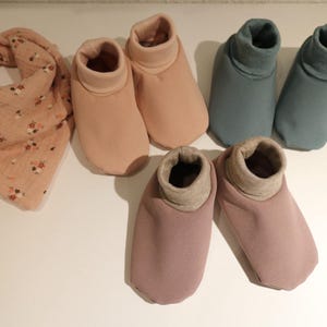 Könnte beinhalten: Eine Auswahl an Babyartikeln, darunter ein rosa Musselin-Lätzchen mit Blumenmuster und mehrere Paare weicher Babyschuhe in Rosa- und Blautönen. Die Schuhe haben ein Design mit gerolltem Saum.