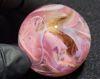 May include: A pink, white, and gold glass marble with swirls and bubbles.