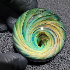 May include: A green, blue, and yellow glass marble with a swirling pattern that creates a vortex effect.