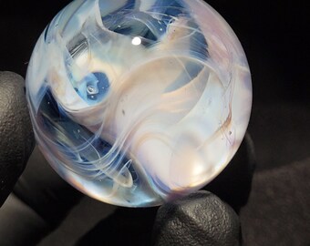 May include: A clear glass marble with swirling patterns of white, blue, and tan. The marble is round and appears to be handmade, with a glossy finish. The background is black, highlighting the marble's colors.