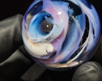 Puede incluir: Una canica de vidrio con colores azul, blanco y morado en espiral. La canica tiene un acabado brillante y parece estar hecha a mano.
