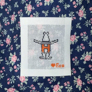 May include: A square, white needlepoint canvas featuring a cartoon figure with outstretched arms and an orange "H" in the center. The figure is black and white. The text "Ree" is below the figure, with a heart symbol.