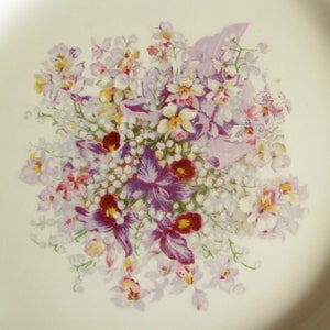 May include: A white ceramic plate with a floral design featuring purple, pink, and yellow flowers. The flowers are arranged in a bouquet and are surrounded by white flowers.