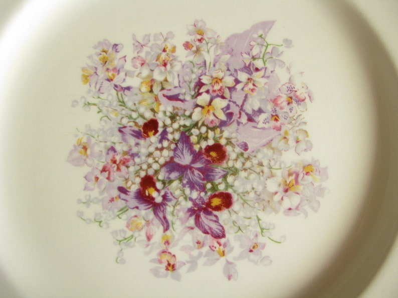 May include: A white ceramic plate with a floral design featuring purple, pink, and yellow flowers. The flowers are arranged in a bouquet and are surrounded by white flowers.
