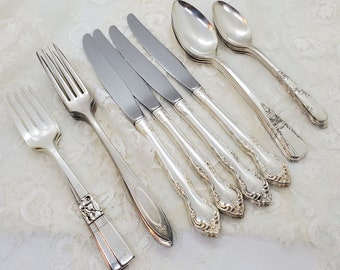 May include: A set of seven silver-plated flatware pieces, including forks and knives, with ornate handles. The flatware is arranged on a white surface.