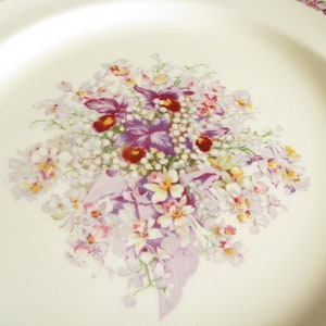 May include: A white ceramic plate with a floral design featuring a bouquet of purple, white, and yellow flowers. The flowers are arranged in a cluster and have a delicate, vintage aesthetic.