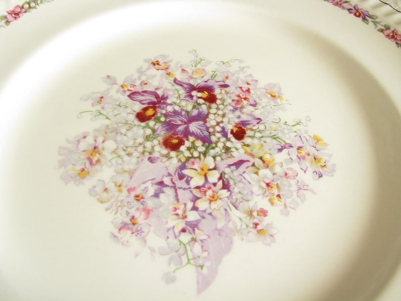 May include: A white ceramic plate with a floral design featuring a bouquet of purple, white, and yellow flowers. The flowers are arranged in a cluster and have a delicate, vintage aesthetic.