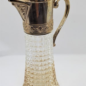 Könnte beinhalten: Eine klare Glaskaraffe mit einem goldfarbenen Metalldeckel und Griff. Die Karaffe hat ein strukturiertes, rautenförmiges Muster.