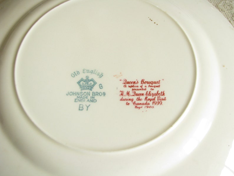 May include: A white ceramic plate with a blue and green design that says "Old English" and "Johnson Bros Made in England By". The plate also has a red inscription that says "Queen's Bouquet A replica of a bouquet presented to H.M. Queen Elizabeth during the Royal Visit to Canada 1939. Reg'd 1940."