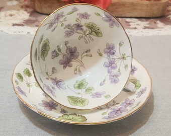 May include: A white teacup and saucer with a floral pattern of purple flowers and green leaves. The cup and saucer have a gold trim.