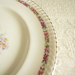 May include: A white ceramic plate with a floral design. The plate has a gold rim and a border of pink and purple flowers. The center of the plate features a cluster of pink and purple flowers.