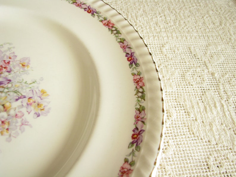 May include: A white ceramic plate with a floral design. The plate has a gold rim and a border of pink and purple flowers. The center of the plate features a cluster of pink and purple flowers.