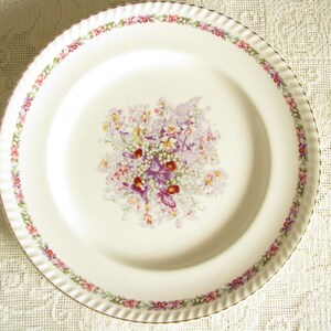 May include: A white ceramic dinner plate with a gold rim and a floral design in shades of pink, purple, and green.