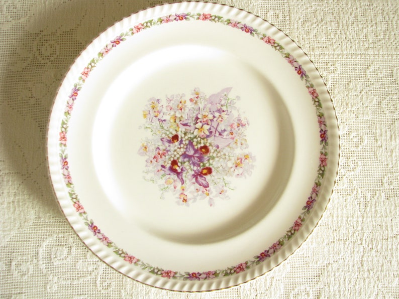 May include: A white ceramic dinner plate with a gold rim and a floral design in shades of pink, purple, and green.