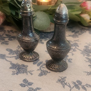 May include: Two silver salt and pepper shakers with ornate designs on a white tablecloth with blue floral pattern.