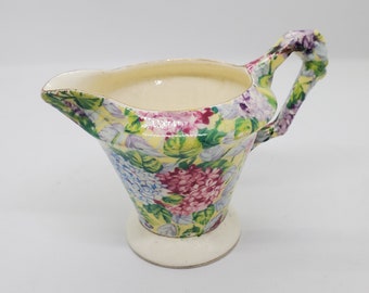 Könnte beinhalten: Ein kleiner, cremefarbener Milchkännchen mit einem Blumenmuster. Der Milchkännchen hat einen Griff und einen breiten Boden. Die Blumen sind rosa, lila, blau und gelb.