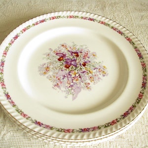 May include: White ceramic dinner plate with a floral design featuring a bouquet of purple, pink, and white flowers with green leaves. The plate has a gold rim and a scalloped edge.