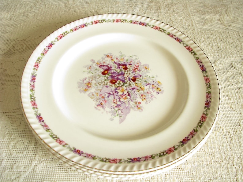 May include: White ceramic dinner plate with a floral design featuring a bouquet of purple, pink, and white flowers with green leaves. The plate has a gold rim and a scalloped edge.