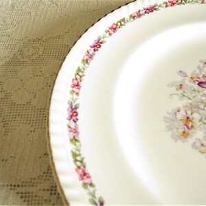 May include: White ceramic plate with a gold rim and a floral design featuring purple, pink, and white flowers. The plate has a scalloped edge.