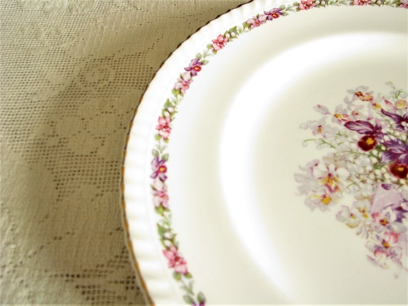 May include: White ceramic plate with a gold rim and a floral design featuring purple, pink, and white flowers. The plate has a scalloped edge.