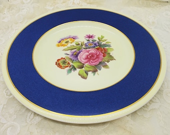 May include: A white ceramic plate with a blue rim and gold trim. The center of the plate features a floral design with pink, yellow, and purple flowers.