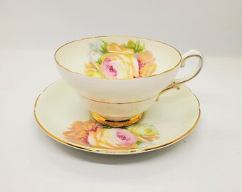 Puede incluir: Una taza de té y un platillo blancos con un diseño floral con rosas rosas y amarillas. La taza y el platillo tienen un borde dorado.