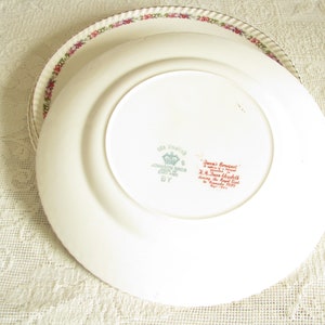 May include: A white ceramic plate with a floral border and a red and green inscription on the bottom. The inscription reads "Made in England, Johnson Bros. England,  'June Bouquet' a design by  J.H.  Town  Meredith during the Royal Visit to Canada, 1939."
